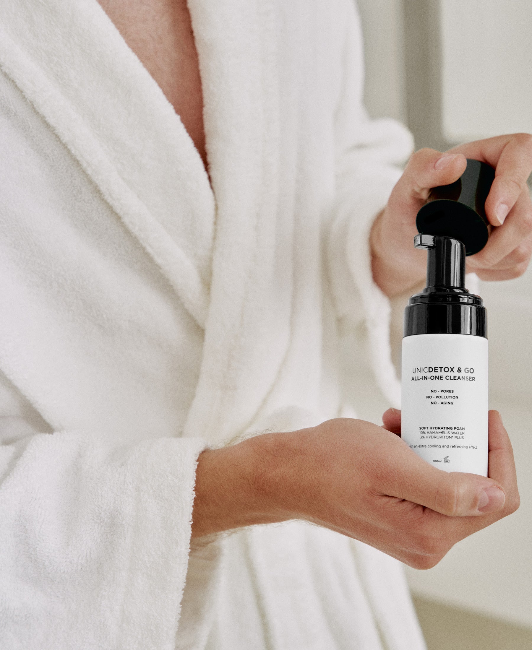 A person in a white bathrobe holds and dispenses foam from a bottle of UNICDETOX & GO All-in-One Cleanser, with the label visible and details slightly blurred.