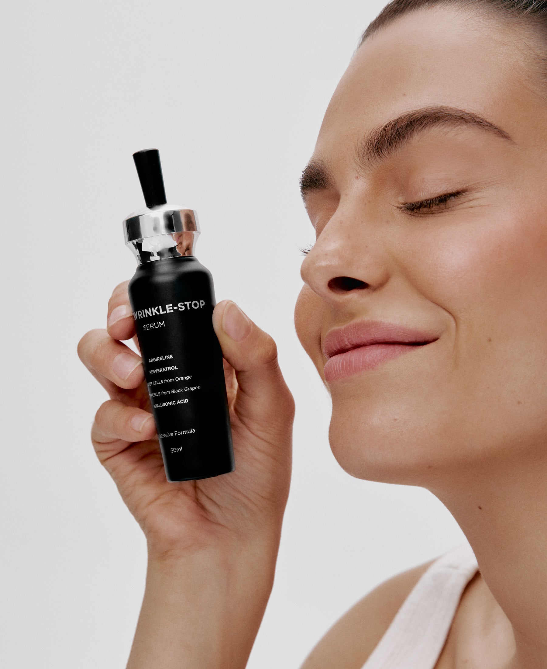 A woman with closed eyes smiles and holds a black bottle labeled "Wrinkle-Stop Serum" near her face, suggesting satisfaction with the skincare product.