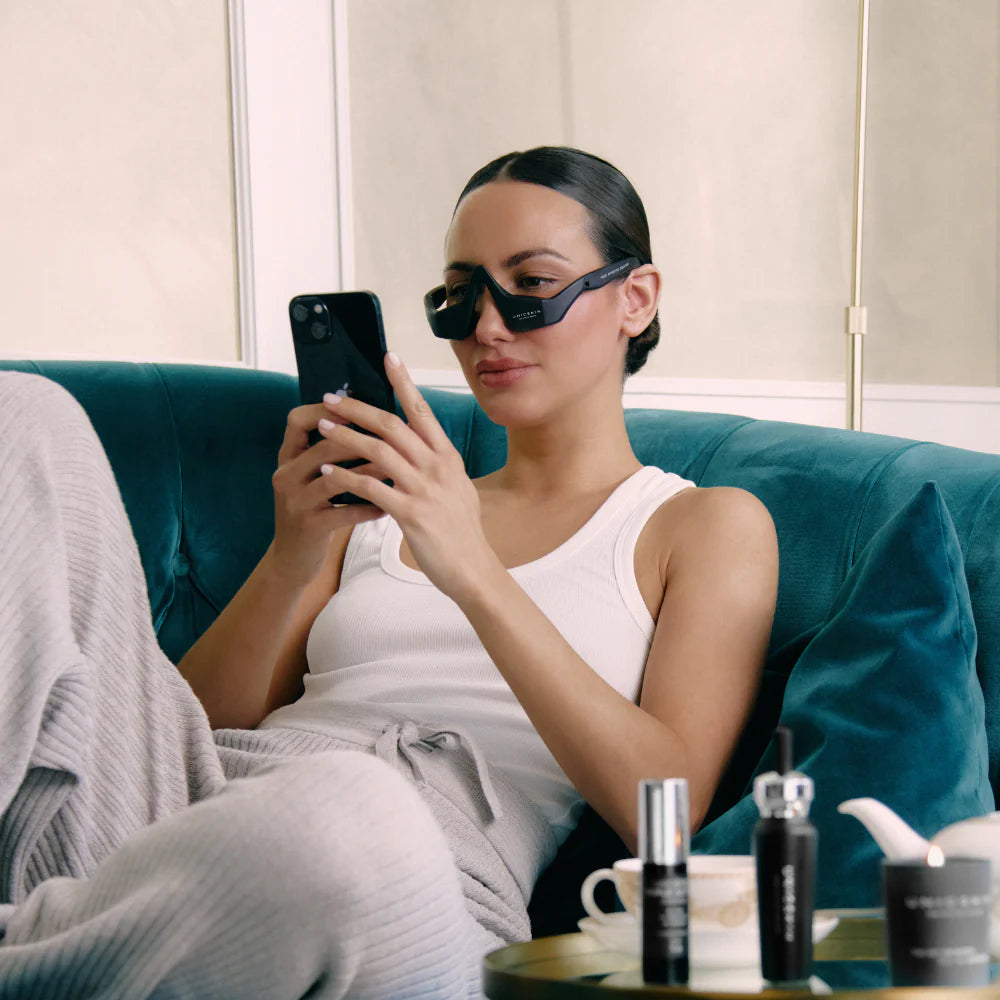 A woman in loungewear relaxes on a teal sofa, wearing a white tank top and grey pants, using the Unic Skin UNICskin UNICLED Eye Mask to soothe tired eyes and reduce puffiness. Skincare products and a teacup sit on the table beside her.