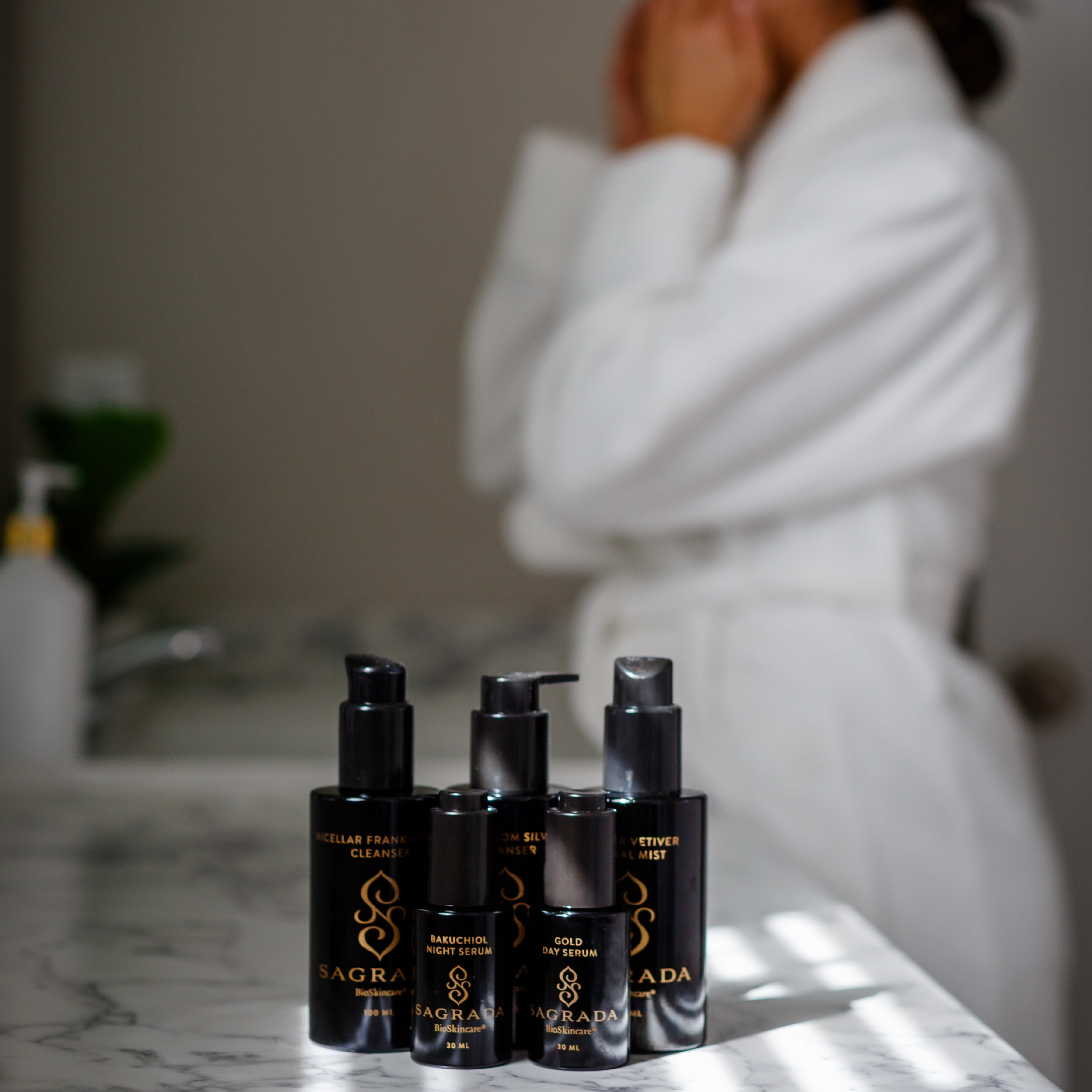 Four black skincare bottles labeled "Sagrada" are on a marble counter, with a person in a white robe blurred in the background, touching their face in a bathroom setting.