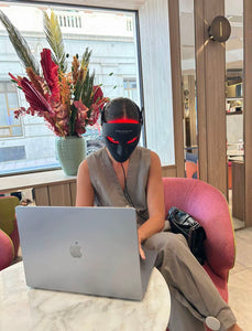 Person sitting at a table with a laptop, wearing a mask, in a cafe setting.