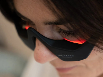 Close-up of a person wearing the Unic Skin UNICskin UNICLED Eye Mask—soothing tired eyes and lifting puffiness with red LED light, which illuminates their eyes and nose against a softly blurred background.