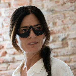A woman with dark braided hair wears the Unic Skin UNICLED Eye Mask—designed to soothe tired eyes and lift puffiness for a refreshed look—paired with a white shirt, standing before a rustic brick wall.