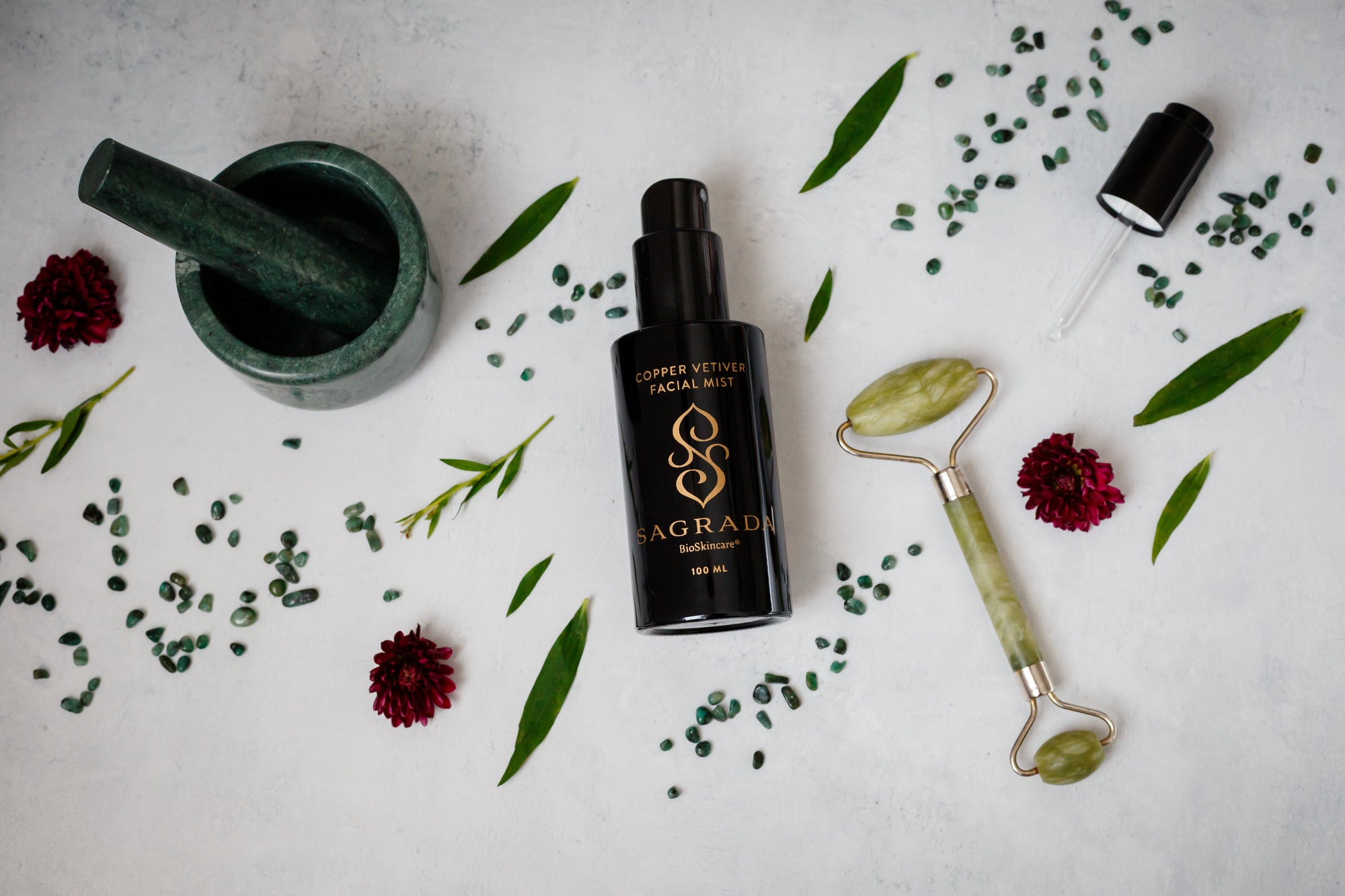 A flat lay of modern skincare items, including a green jade roller, Sagrada BioSkincare’s Sagrada Copper Vetiver Face Mist - Awaken, Balance, & Renew, a small mortar and pestle, green leaves, red flowers, and green beads on a white background.