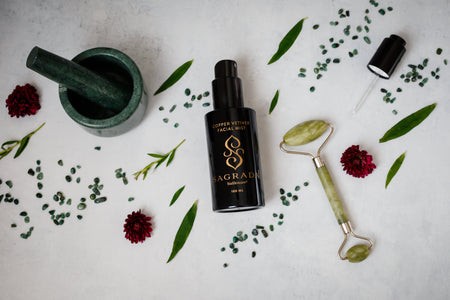 A flat lay of modern skincare items, including a green jade roller, Sagrada BioSkincare’s Sagrada Copper Vetiver Face Mist - Awaken, Balance, & Renew, a small mortar and pestle, green leaves, red flowers, and green beads on a white background.