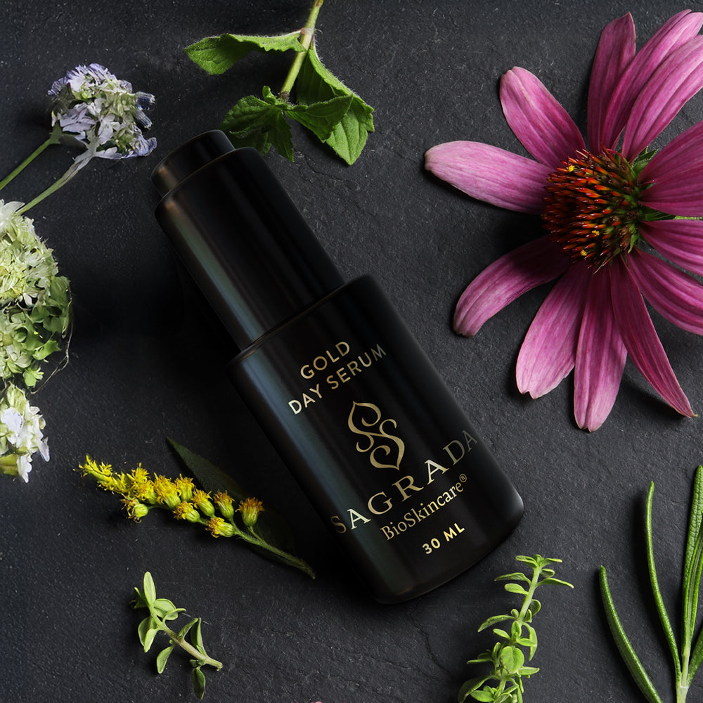 A black bottle labeled “Sagrada Day Gold Serum - Radiance Boosting Elixir” by Sagrada BioSkincare rests on a dark stone surface amid colorful flowers, reflecting its blend of colloidal gold and Helichrysum essential oil.
