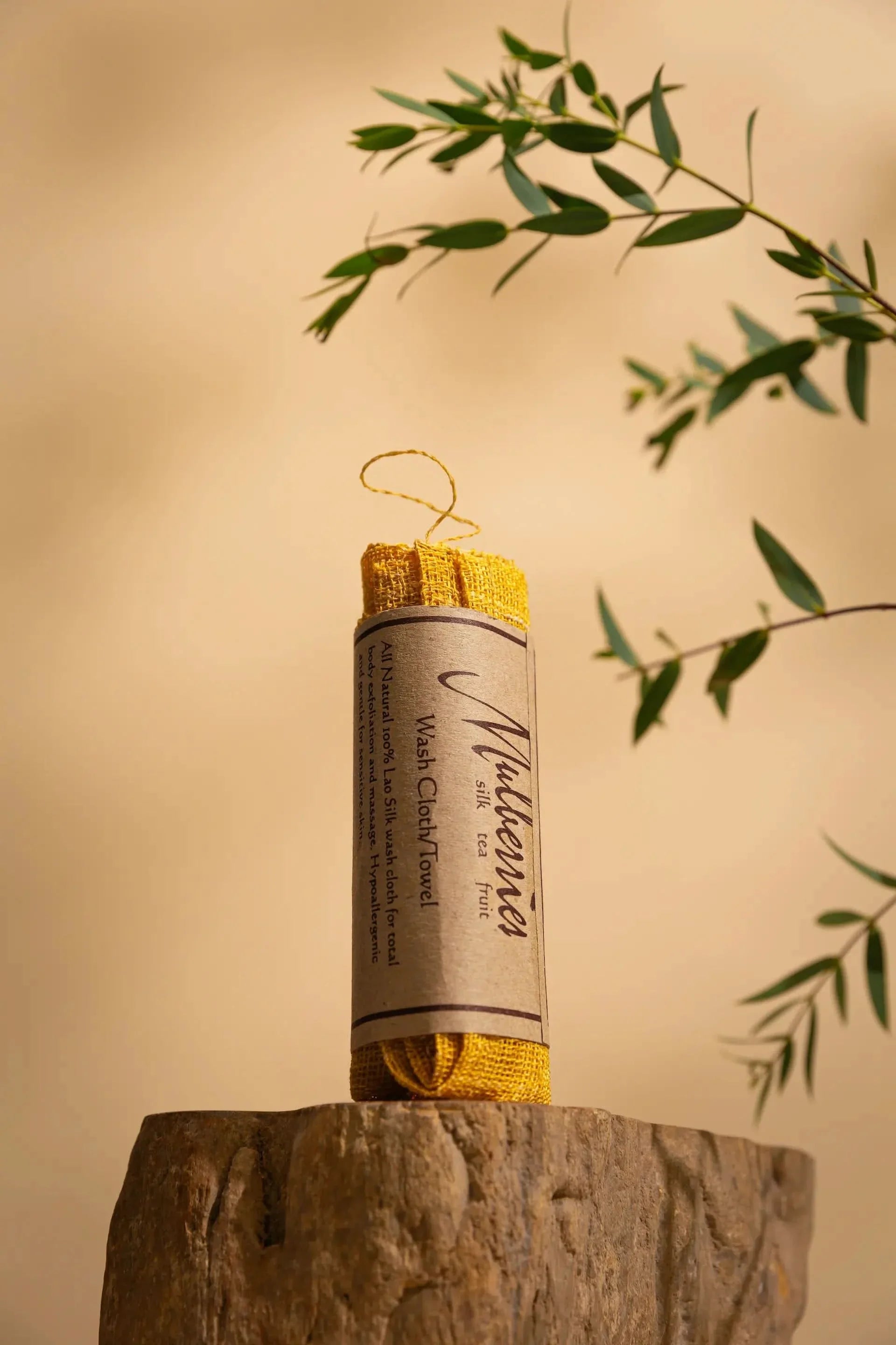 The Sagrada Raw Silk Face Cloth by Sagrada BioSkincare, designed to buff away dry skin and encourage healthy circulation for luminous skin, stands rolled on wood with green leaves framing it against a beige background.