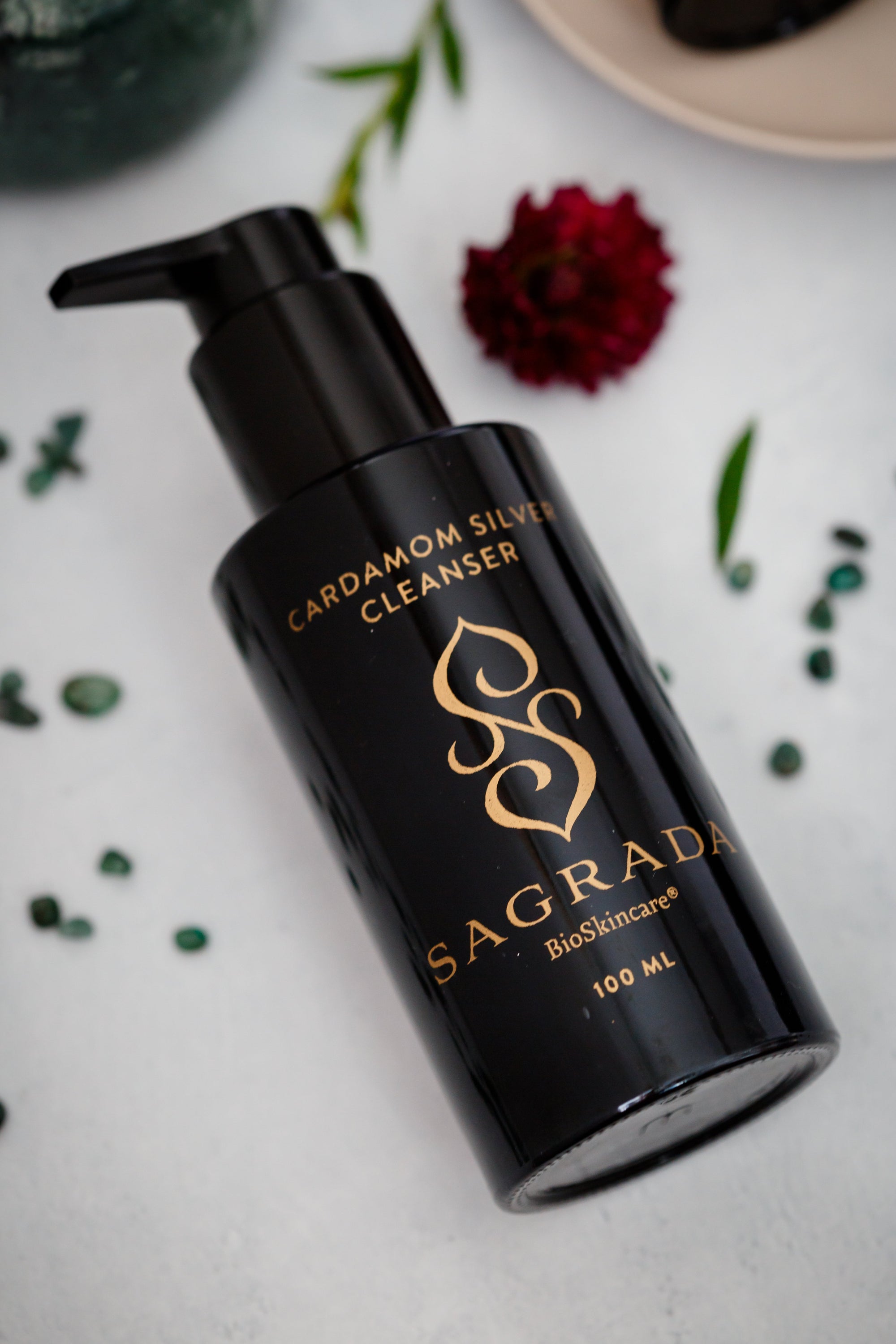 A black bottle of Sagrada Cardamom Silver Cleanser by Sagrada BioSkincare with a pump lies on a white surface, decorated with green leaves and a red flower, emphasizing antioxidant-rich olive oil for deep purification.