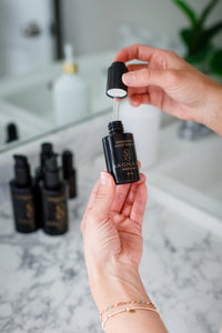 A person holds a black dropper bottle labeled "Sagrada Night Bakuchiol Serum - Gentle Alternative To Retinol" by Sagrada BioSkincare over a marble bathroom counter, with other black bottles and a white soap dispenser in the background.