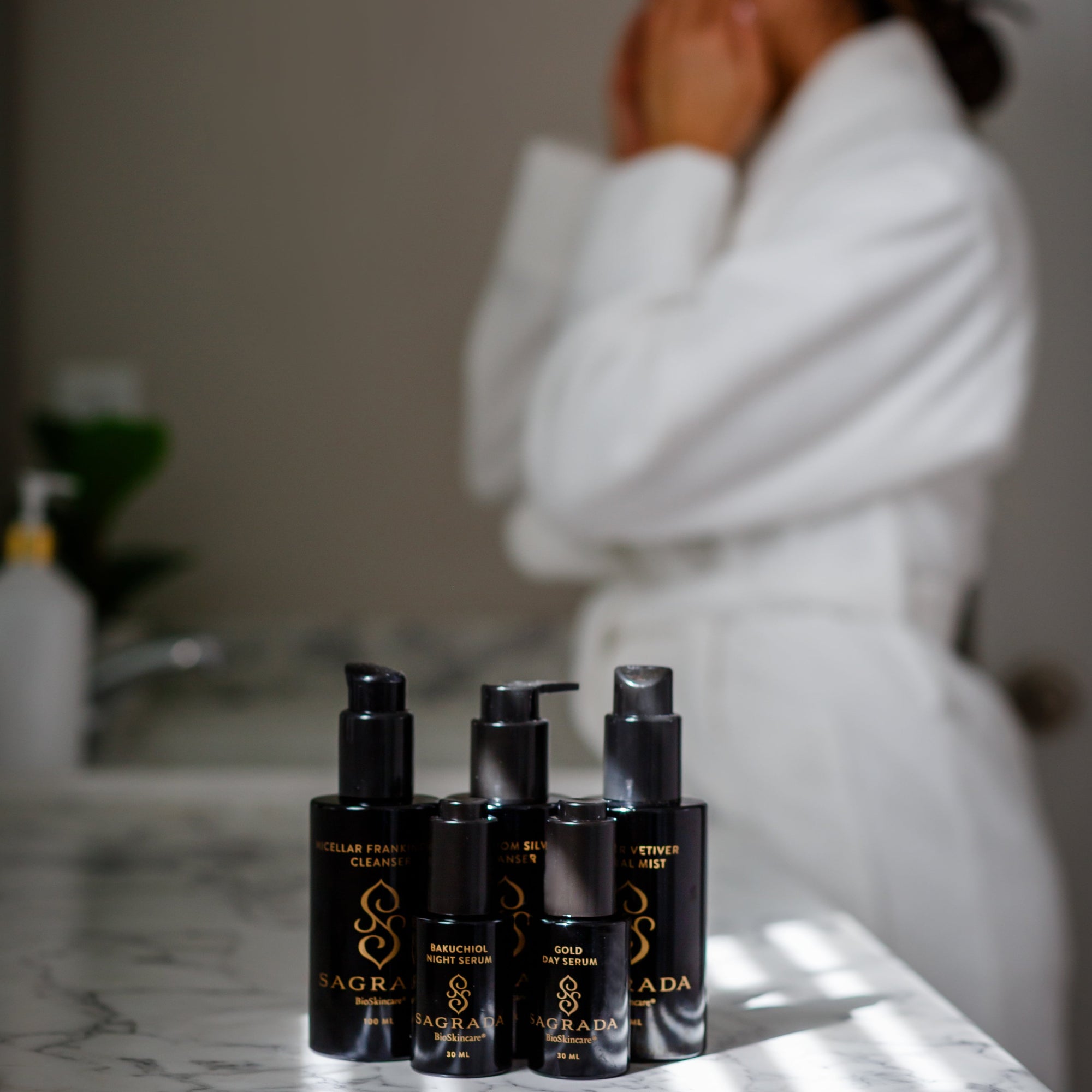 Four black bottles of Sagrada BioSkincare rest on a marble counter, with the Cardamom Silver Cleanser—For Those Who Seek Deep Purification—featured. In the blurred background, a person in a white bathrobe applies product to their face.