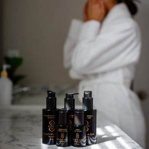 Four black bottles of Sagrada BioSkincare rest on a marble counter, with the Cardamom Silver Cleanser—For Those Who Seek Deep Purification—featured. In the blurred background, a person in a white bathrobe applies product to their face.
