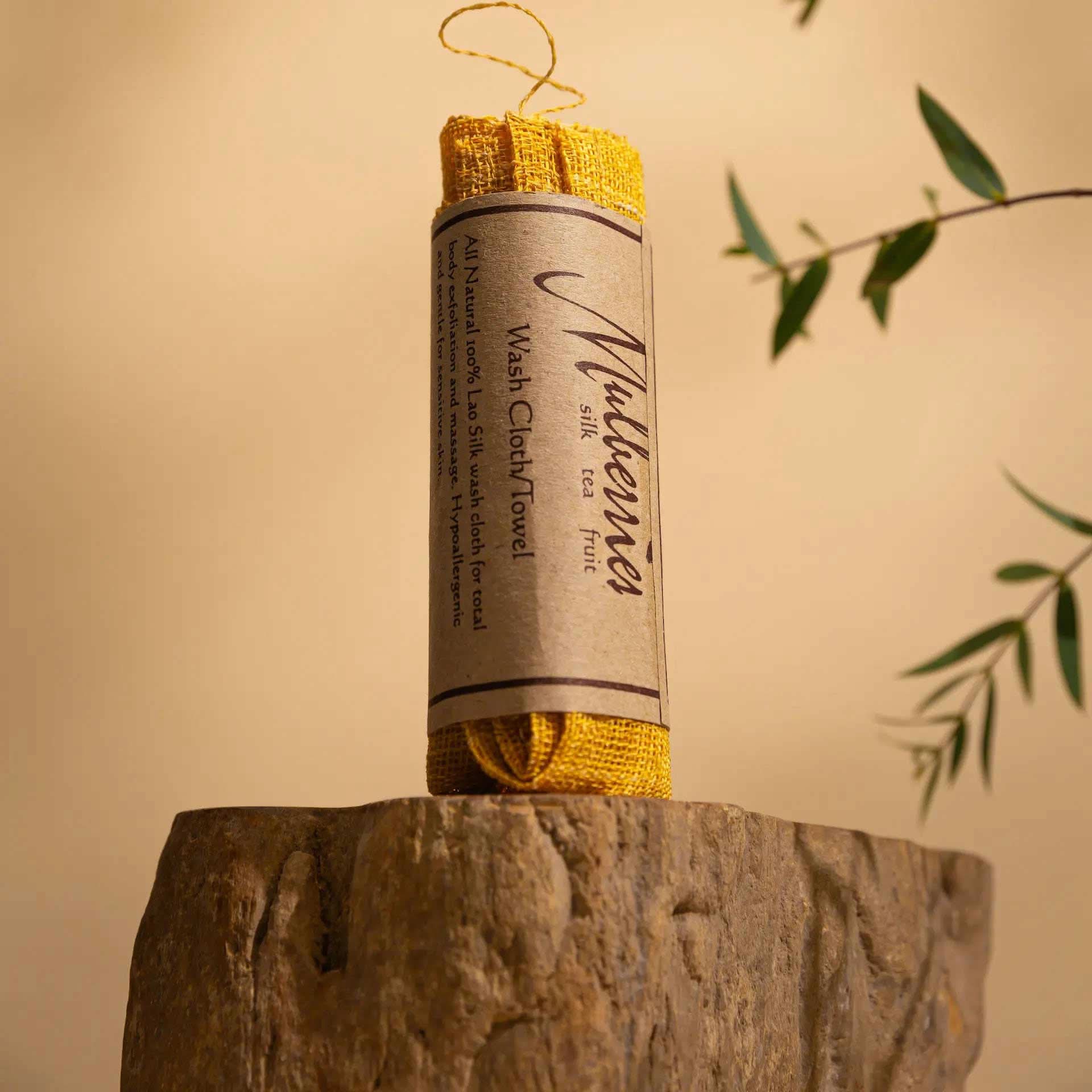 The Sagrada Raw Silk Face Cloth by Sagrada BioSkincare, wrapped in a brown label, stands on a textured stone surface with green leaves and a beige background—buffing away dry skin for radiant, healthy circulation.