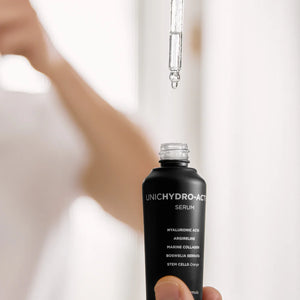 A hand holds a dropper above an open black bottle labeled "UNICHYDRO-ACTIVE" by Adverto Beauty, an intensive hydrating, soothing, and anti-aging facial serum—ideal for dehydrated and sensitive skin. A drop is about to fall into the bottle.