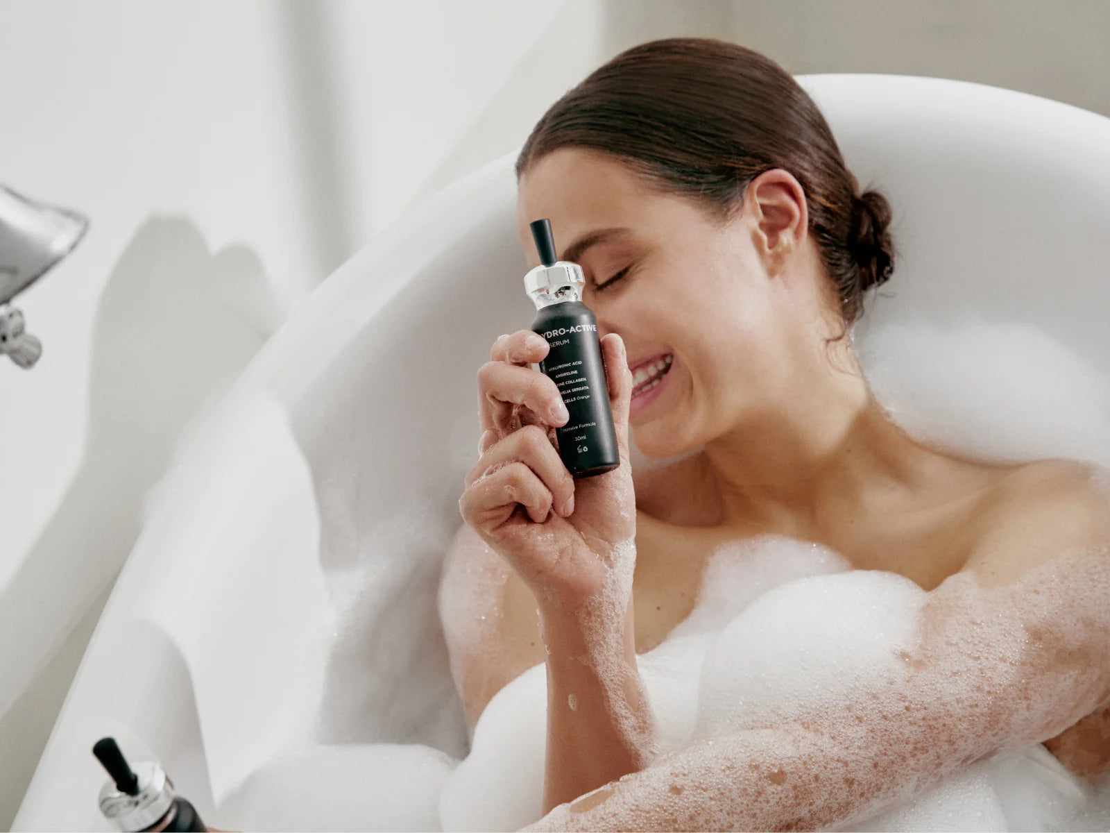 A woman relaxes in a bubble bath, smiling with her eyes closed as she holds Adverto Beauty's UNICHYDRO-ACTIVE Intensive Hydrating, Soothing & Anti-Aging Facial Serum close to her face, enjoying gentle care for sensitive skin.