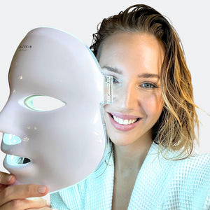 A smiling woman in a white robe uses the UNICskin Korean LED Mask by Unic Skin, holding the glowing device next to her face to reveal its advanced LED therapy for acne, hyperpigmentation, and aging as part of her skincare routine.