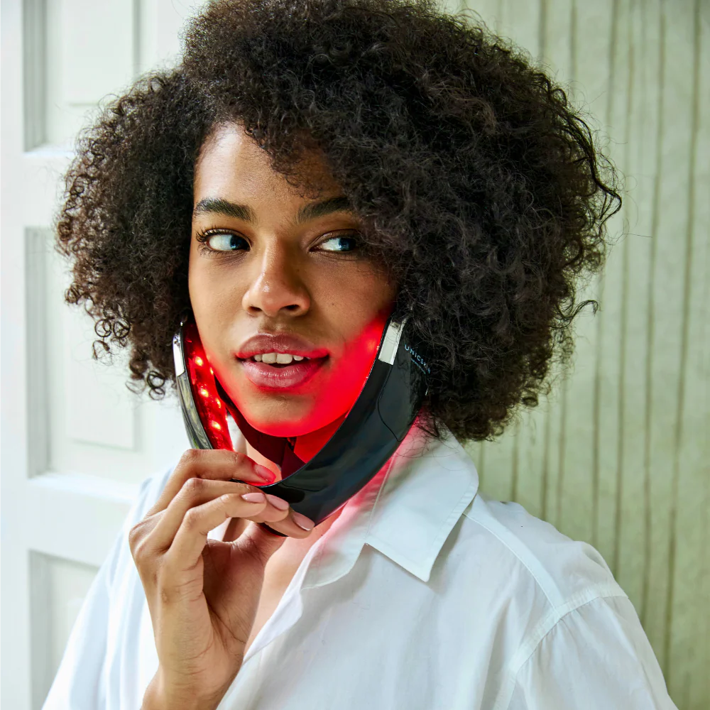 A person with curly hair uses the Unic Skin UNICskin UNICLED Neck & Chin Device—a black LED light therapy mask emitting red lights to firm and tighten skin for a contoured neck and jawline. They wear a white shirt, indoors near a white door and textured wall.