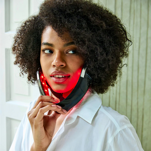 A person with curly hair uses the Unic Skin UNICskin UNICLED Neck & Chin Device—a black LED light therapy mask emitting red lights to firm and tighten skin for a contoured neck and jawline. They wear a white shirt, indoors near a white door and textured wall.