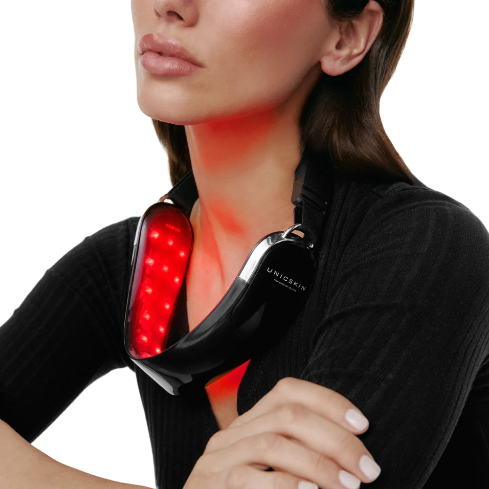 A woman wears the Unic Skin UNICLED Neck & Chin Device, its red LED lights glowing around her neck as she rests her arms on a table and looks upward. The device is designed to firm, tighten, and contour the neck and jawline.