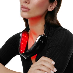 A woman wears the Unic Skin UNICLED Neck & Chin Device, its red LED lights glowing around her neck as she rests her arms on a table and looks upward. The device is designed to firm, tighten, and contour the neck and jawline.