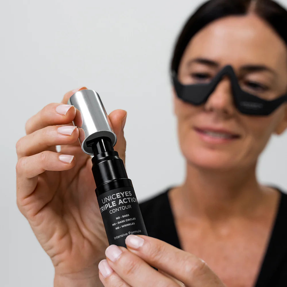 A woman wearing the black UNICskin UNICLED Eye Mask holds and opens a bottle labeled "UNICEYES TRIPLE ACTION CONTOUR" by Unic Skin, softly smiling.