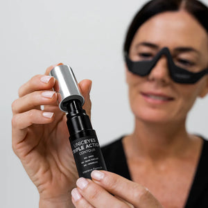 A woman wearing the black UNICskin UNICLED Eye Mask holds and opens a bottle labeled "UNICEYES TRIPLE ACTION CONTOUR" by Unic Skin, softly smiling.