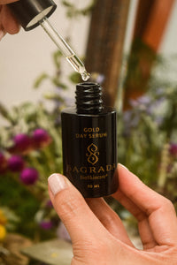 A hand holds a black bottle of Sagrada BioSkincare’s Sagrada Day Gold Serum - Radiance Boosting Elixir while another hand uses a dropper to dispense the gold-infused serum, with blurred flowers in the background.
