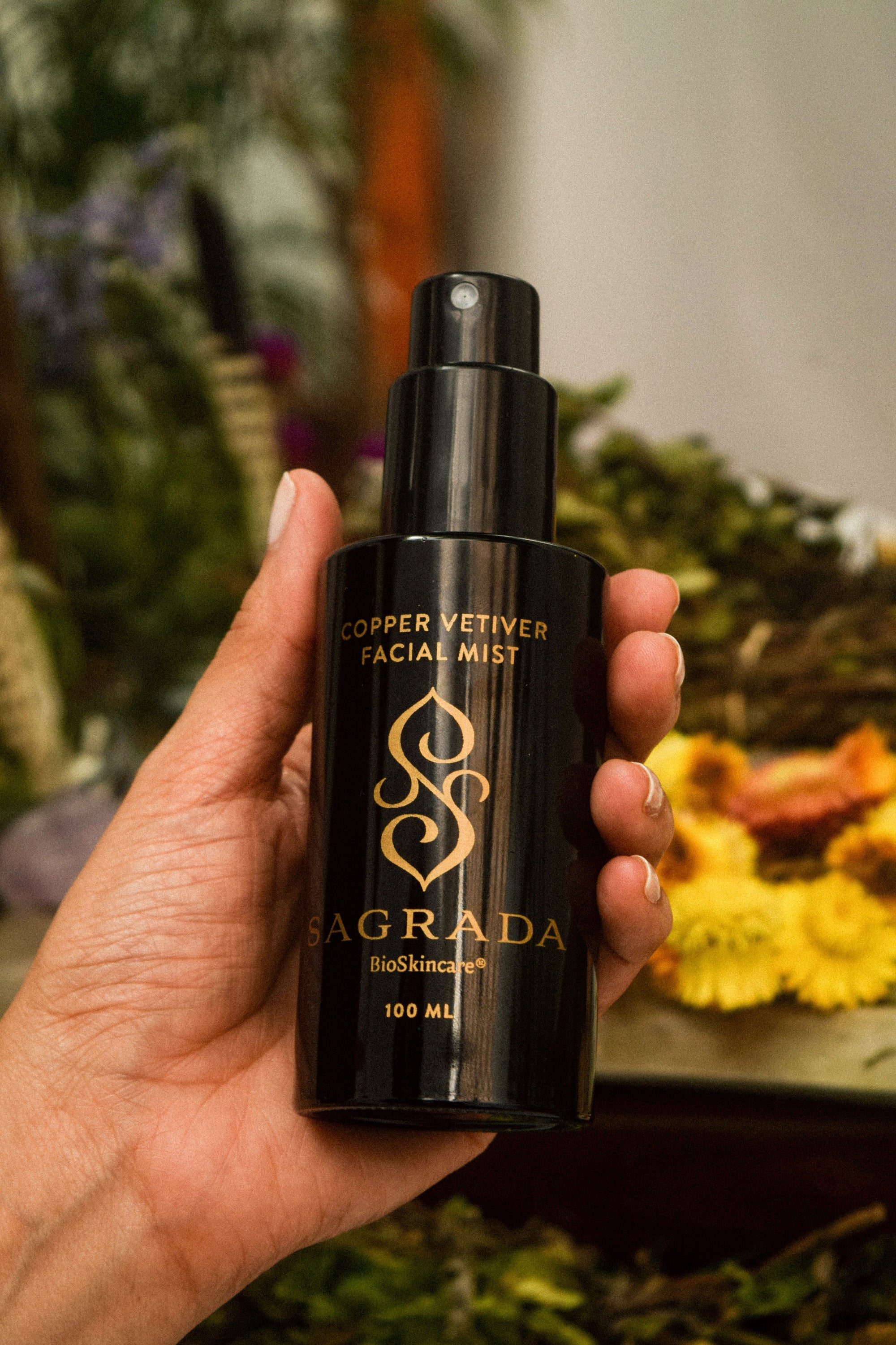 A hand holds a black bottle labeled "Sagrada Copper Vetiver Face Mist – Awaken, Balance, & Renew" by Sagrada BioSkincare, with green leaves and yellow flowers softly blurred in the background.