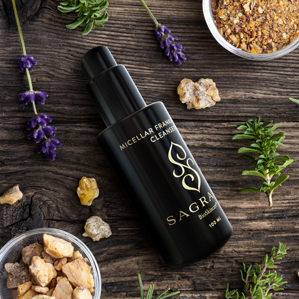 A black bottle of Sagrada BioSkincare’s Sagrada Micellar Frankincense Cleanser – Deep Cleanser For Delicate Skin sits on rustic wood, surrounded by lavender, rosemary, resin chunks, and herbal mixtures—showcasing gentle micellar technology.