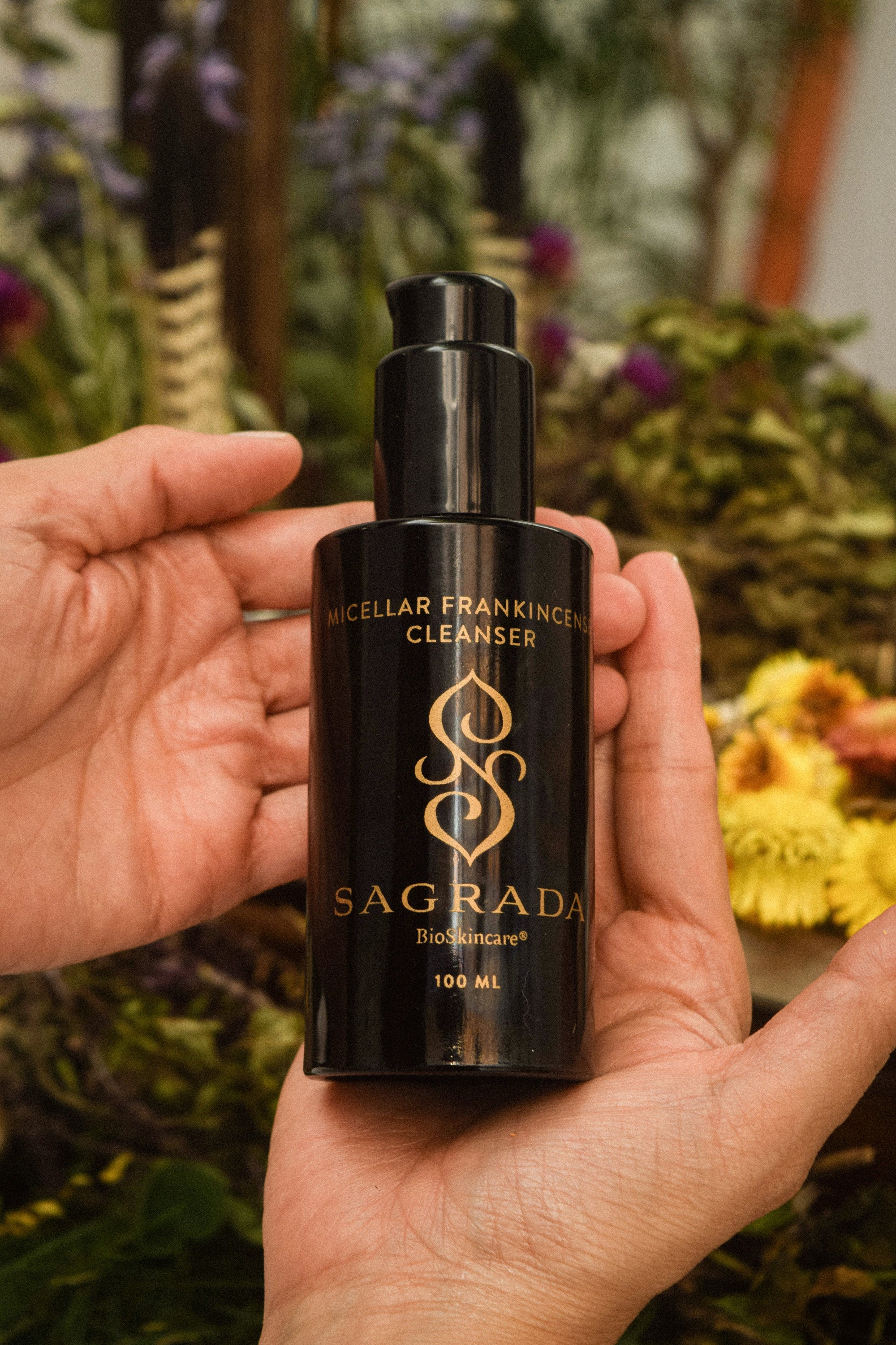 A hand holds a dark bottle labeled "Sagrada BioSkincare Micellar Frankincense Cleanser - Deep Cleanser For Delicate Skin" with a pump dispenser; blurred flowers and plants in the background highlight its gentle yet powerful cleansing.