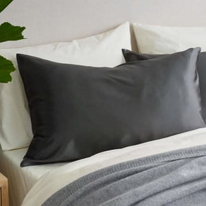 A neatly made bed with white sheets, a gray blanket, and two dark gray Sagrada Mulberry Silk Pillowcases by Sagrada BioSkincare pampers your skin and hair, set against a light-colored wall with part of a green plant visible on the left.