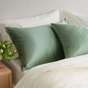 Two light green Sagrada Mulberry Silk Pillowcases from Sagrada BioSkincare rest on a white bed, with a leafy plant nearby, highlighting their luxurious, skin- and hair-pampering, thermoregulating benefits.