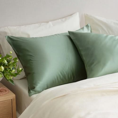 Two light green Sagrada Mulberry Silk Pillowcases from Sagrada BioSkincare rest on a white bed, with a leafy plant nearby, highlighting their luxurious, skin- and hair-pampering, thermoregulating benefits.